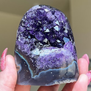 May include: A polished amethyst geode specimen featuring a deep purple crystalline cluster at the top, transitioning into a smooth, banded gray and blue agate base with visible natural mineral layers and a rounded, dome-like shape.