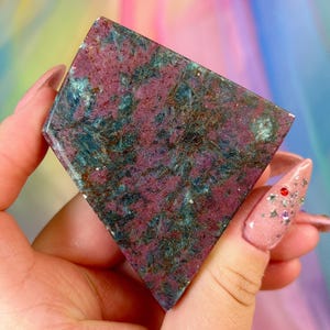 May include: A polished, shield-shaped slab of ruby kyanite mineral featuring a mottled texture with deep magenta, dark blue, and metallic bronze inclusions. The stone has a flat, smooth surface with a distinct geometric cut and natural crystalline patterns.