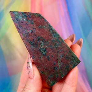 May include: A polished, wedge-shaped mineral specimen featuring a deep burgundy red base color with scattered inclusions of dark green and metallic blue mineral flecks throughout the surface, displaying a smooth, flat, and angled top edge.
