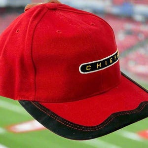 May include: A red baseball cap with a black brim and the word "CHIEFS" embroidered in white on a black rectangle. The cap is set against a blurred background of a football field.