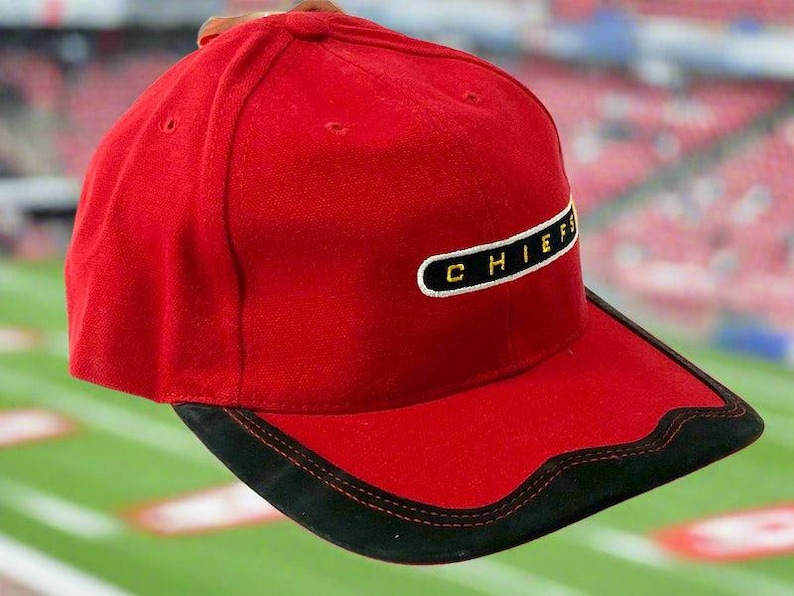 May include: A red baseball cap with a black brim and the word "CHIEFS" embroidered in white on a black rectangle. The cap is set against a blurred background of a football field.