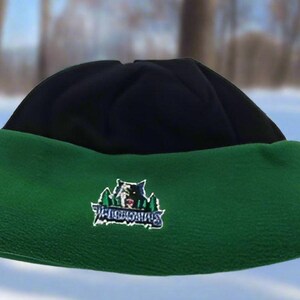 May include: A black and green beanie hat with a folded brim. The green brim features an embroidered logo with a bear and trees. The hat is designed for cold weather and outdoor activities.