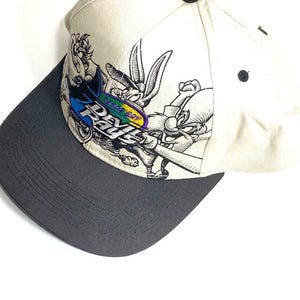 May include: Cream-colored baseball cap with a dark gray brim. The cap features a cartoon graphic of Bugs Bunny with the text "Day Rave" in blue, purple, and green.