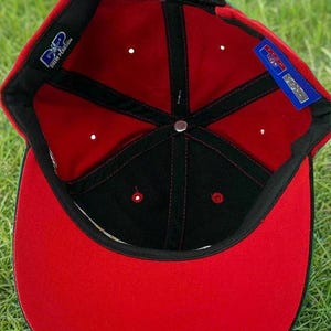 May include: A red and black baseball cap with a curved brim. The interior is red with black stitching and a black band. The cap has a green accent on the back and a small tag with text. The cap is sitting on green grass.