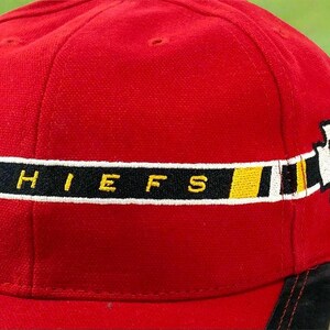 May include: A red baseball cap with the word "CHIEFS" embroidered in yellow letters on a black stripe. The cap also features a white arrowhead logo with a red "K" and a yellow stripe.