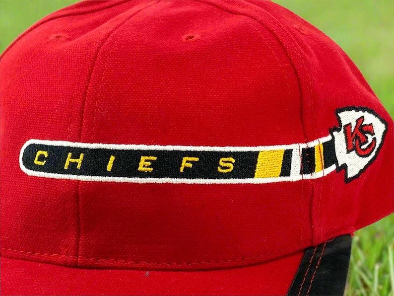 May include: A red baseball cap with the word "CHIEFS" embroidered in yellow letters on a black stripe. The cap also features a white arrowhead logo with a red "K" and a yellow stripe.