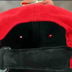 May include: A red baseball cap with a black interior and a black adjustable strap. The cap has a curved brim and a tag attached to the top. The cap is made of a textured fabric.