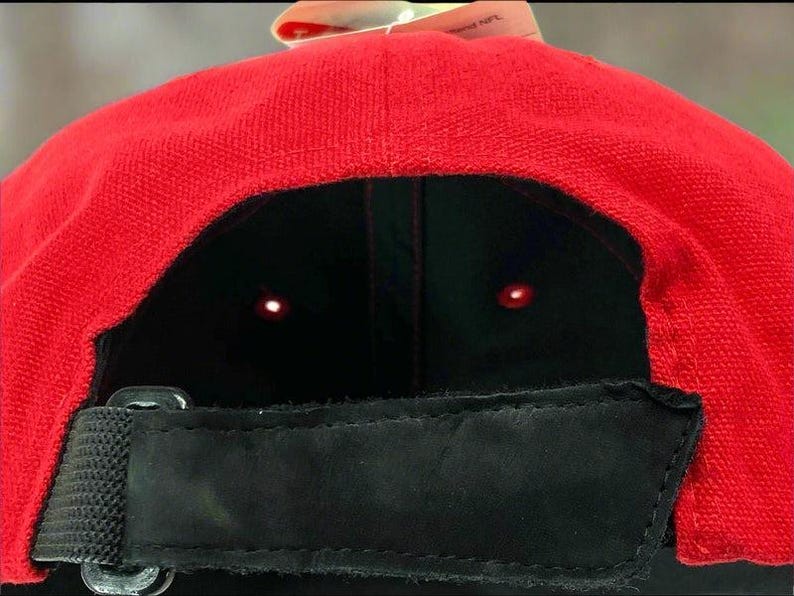 May include: A red baseball cap with a black interior and a black adjustable strap. The cap has a curved brim and a tag attached to the top. The cap is made of a textured fabric.