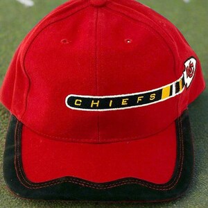 May include: A red baseball cap with a black brim and the word "CHIEFS" in white letters. The cap features a logo of a hockey stick and a feather. The hat is made of a soft material.