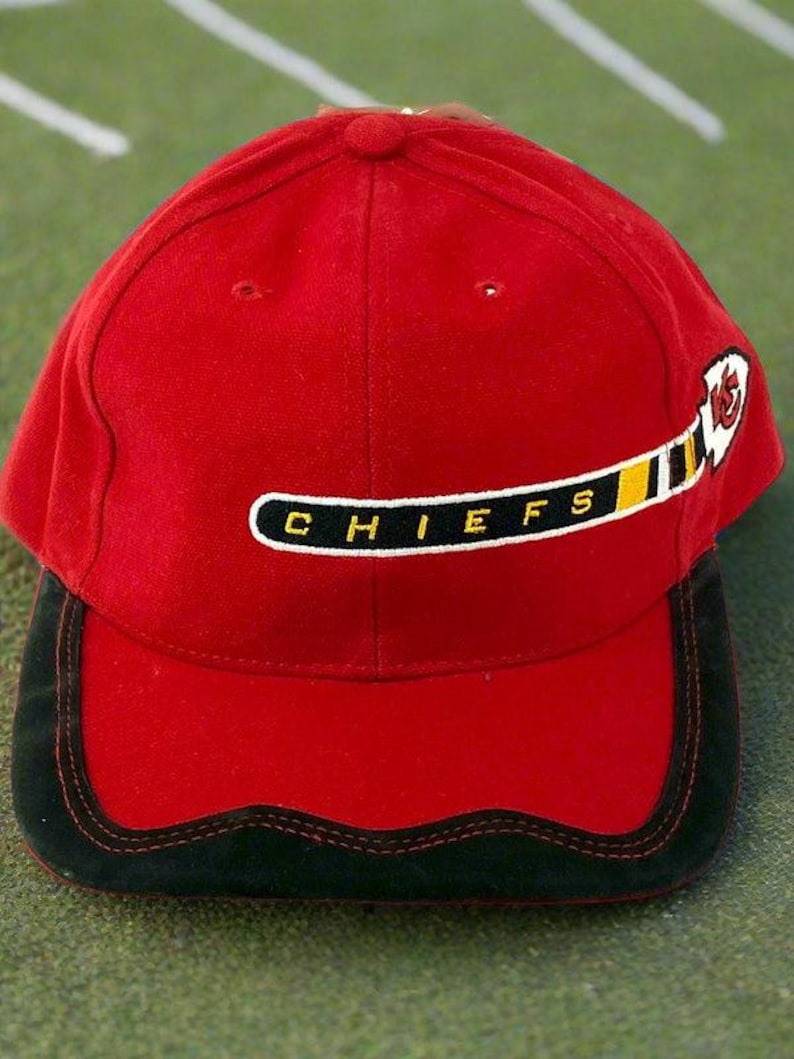May include: A red baseball cap with a black brim and the word "CHIEFS" in white letters. The cap features a logo of a hockey stick and a feather. The hat is made of a soft material.