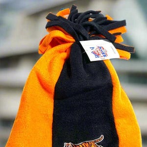 May include: A fleece beanie in orange and black, featuring a tiger logo. The hat has a tassel on top and a tag attached. The design is a color block style with orange on the sides and black in the center.