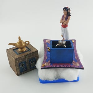 May include: A miniature scene featuring a gold lamp atop a brown box with a key and lock design. A blue box with a ring sits on a magic carpet, with a figurine of a character from Aladdin standing nearby.