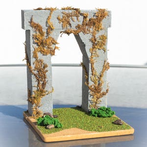 May include: A miniature stone archway covered in brown vines, set on a small patch of green grass with brown sand and small green bushes.