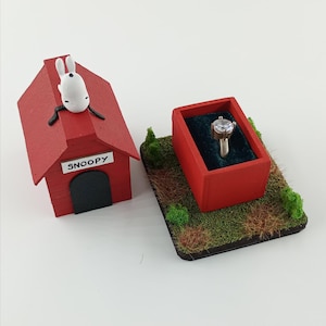 May include: A red Snoopy dog house with a white Snoopy figure on top, next to a red ring box with a diamond ring inside. The ring box sits on a small patch of grass with miniature trees.