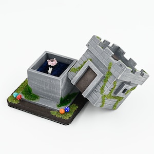 May include: A gray stone ring box shaped like a castle with a pink gemstone ring inside. The box is decorated with green moss and has a small blue, orange, and red decorative element on the base.