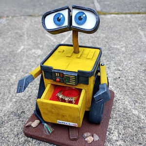 May include: A yellow robot with a black drawer that is open, revealing a red interior and a gold ring. The robot has blue eyes and a black antenna. The robot is sitting on a brown base with the word "WALL-E" printed on it.