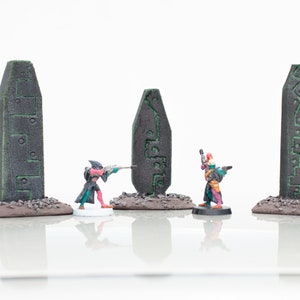 Puede incluir: Tres figuras en miniatura se encuentran frente a tres pilares de piedra gris oscuro y altos con marcas verdes. Las figuras visten ropa colorida y sostienen armas. Los pilares están sobre una base marrón.