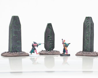 May include: Three miniature figures stand in front of three tall, dark gray stone pillars with green markings. The figures are wearing colorful clothing and holding weapons. The pillars are on a brown base.