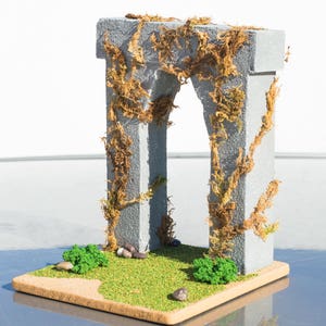 May include: A miniature stone archway covered in brown vines, set on a base with green grass and small stones.
