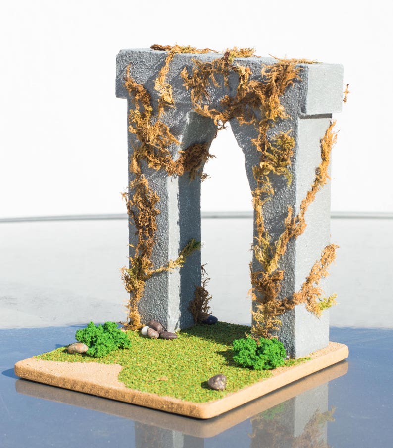 May include: A miniature stone archway covered in brown vines, set on a base with green grass and small stones.