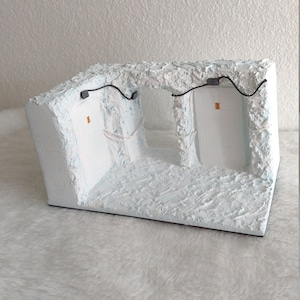 May include: A white, textured, rectangular diorama with two white rectangular openings. The openings are framed by white, textured walls and have black wires running through them. The diorama is sitting on a white, fluffy surface.