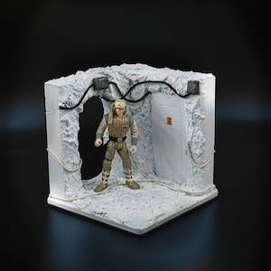 Könnte beinhalten: Ein weißes, schneebedecktes Diorama mit einer Star Wars Figur von Luke Skywalker in einem beigefarbenen Overall und einem schwarzen Helm, der im Türrahmen steht. Das Diorama hat eine weiße Tür mit einem schwarzen Warnschild und schwarzen Drähten, die an den Wänden entlanglaufen.