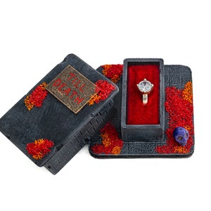 May include: A black ring box with a red velvet interior and a diamond ring. The box is decorated with a skull and the words "Till Death".