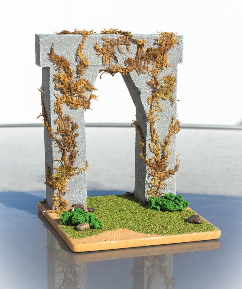 May include: A miniature stone archway covered in brown vines, set on a small patch of green grass with brown sand and small green bushes.
