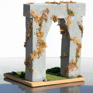 May include: A miniature stone archway covered in brown and green moss. The archway is set on a small, brown base with green grass and small stones.