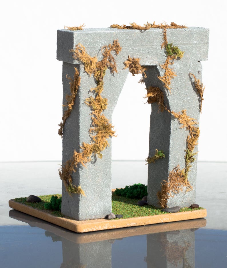 May include: A miniature stone archway covered in brown and green moss. The archway is set on a small, brown base with green grass and small stones.