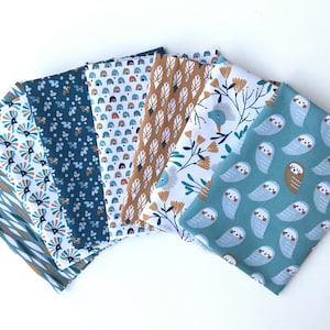 Op de afbeelding: Een verzameling gevouwen stoffen met verschillende patronen. Ontwerpen omvatten geometrische vormen, bloemstukken en grillige uilen in tinten blauw, bruin en wit. De stoffen zijn in een waaier gerangschikt.