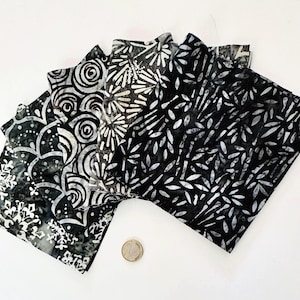 Op de afbeelding: Een verzameling van vijf gevouwen stoffen vierkanten met zwart-witte batikpatronen, waaronder sneeuwvlok-, spiraal- en bladmotieven. Een kleine ronde munt is onderaan in het midden geplaatst voor schaal, met een diameter van 2,3 cm.