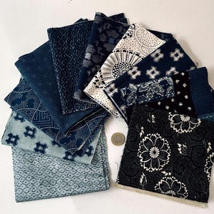 May include: An assortment of folded fabric squares in shades of indigo blue. The patterns include floral, geometric, and abstract designs. Some fabrics are white with blue patterns. A coin is visible for scale.