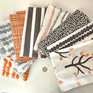 Puede incluir: Una colección de cuadrados de tela doblados con varios patrones, incluyendo hojas naranjas, círculos grises, rayas marrones y puntos blancos. Las telas están dispuestas en forma de abanico, mostrando los diferentes diseños y colores.