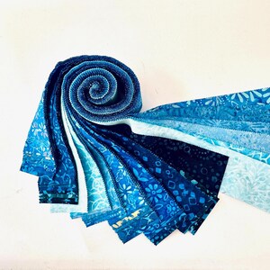 Op de afbeelding: Een rol stofstroken met een verloop van blauwtinten. De collectie bevat diverse patronen zoals bloemmotieven, geometrische vormen en abstracte batikontwerpen, gerangschikt in een spiraal die uitwaaiert in individuele stroken in verschillende blauwtinten.