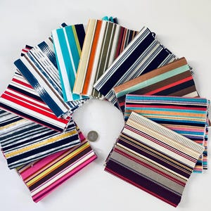 Op de afbeelding: Een verzameling rechthoekige stoffen stalen gerangschikt in een cirkelvormig waaierpatroon. De stalen zijn voorzien van diverse streeppatronen in kleuren zoals turquoise, oranje, marineblauw, rood, roze, geel en wit. Een kleine zilveren munt ligt in het midden.