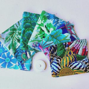 Könnte beinhalten: Eine Sammlung gefalteter Stoffquadrate mit lebendigen Blumen- und Muschelmustern in Blau-, Grün-, Gelb- und Rosatönen. Ein kleines weißes, kreisförmiges Maßband liegt zur Größenbestimmung in der Mitte des Stoffstapels.