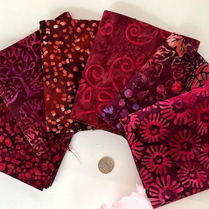 May include: A collection of six folded fabric squares arranged in a fan shape, featuring various batik patterns in shades of deep red, burgundy, and magenta. A small circular coin is placed in the center for scale, measuring 23 millimeters in diameter.