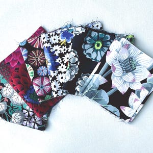 以下が含まれることがあります： さまざまな花柄と抽象柄の5つの正方形の布のコレクション。生地は、黒、赤、青、白など、さまざまな色で、詳細な植物や幾何学模様が特徴です。端は未完成です。