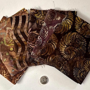 Pode incluir: Uma coleção de cinco amostras de tecido em tons de castanho, bronze e roxo escuro com padrões estilo batik, incluindo espirais, ondas e formas orgânicas. Uma moeda de cor prateada está colocada sob o tecido como referência de escala.