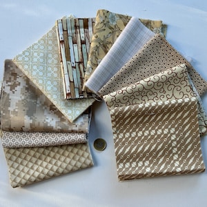 Könnte beinhalten: Ein Stapel von acht Stoffquadraten in verschiedenen Beige- und Brauntönen. Die Stoffe zeigen geometrische Muster, florale Muster und abstrakte Drucke.