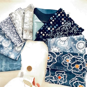 May include: A collection of ten fabric squares arranged in a fan shape, featuring various blue, grey, and white patterns including celestial suns, moons, stars, floral motifs, script text, and abstract dots, with a small circular coin for scale.