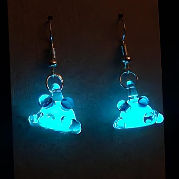May include: A pair of glowing blue glass earrings. Each earring features a unique shape with rounded edges and small, decorative glass beads. The earrings hang from silver-colored fish hook ear wires.
