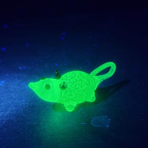 Puede incluir: Un objeto con forma de ratón verde brillante con una cola larga y curvada. El ratón tiene un cuerpo texturizado y ojos pequeños y redondos. El fondo es azul oscuro con motas azul claro dispersas, creando un efecto estrellado.