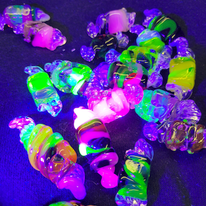May include: A collection of colourful glass beads, some with swirls and others with a smooth finish, glow brightly under a black light. The beads are a mix of green, pink, yellow, and black.