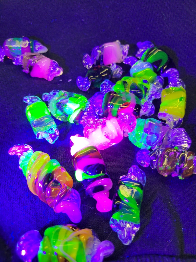 May include: A collection of colourful glass beads, some with swirls and others with a smooth finish, glow brightly under a black light. The beads are a mix of green, pink, yellow, and black.