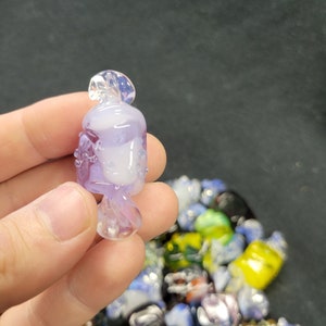 May include: A small, hand-blown glass candy charm in a purple and white swirl pattern. The charm is shaped like a wrapped candy and has a clear glass stem.
