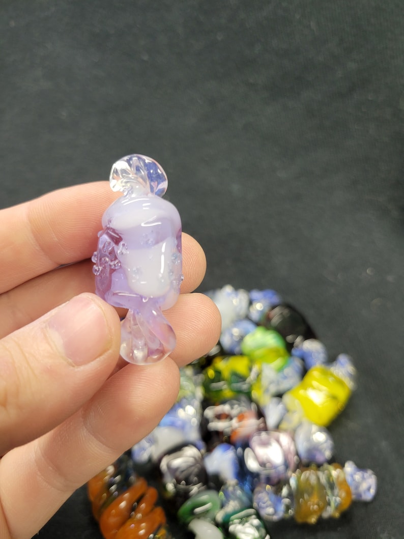 May include: A small, hand-blown glass candy charm in a purple and white swirl pattern. The charm is shaped like a wrapped candy and has a clear glass stem.