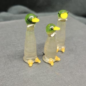 Könnte beinhalten: Drei Glas-Entenfiguren mit grünen Köpfen, gelben Schnäbeln und weißen Augen. Die Enten stehen auf einer grauen Oberfläche.