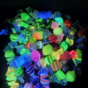 Op de afbeelding: Een verzameling van talloze glazen snoepsculpturen in diverse vormen en maten, voorzien van levendige, veelkleurige patronen die oplichten onder ultraviolet licht, waaronder tinten neon-groen, felroze, elektrisch blauw en oranje.